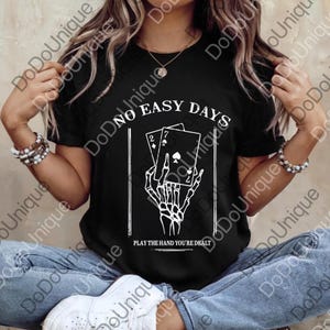 May include: Black t-shirt with the text "NO EASY DAYS" and "PLAY THE HAND YOU'RE DEALT". The graphic features a skeleton hand holding playing cards. The cards show a 2 of diamonds and a 7 of spades.