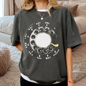 Puede incluir: Camiseta gris oscuro con un diseño de disco telefónico rotatorio vintage. El disco es blanco con números y letras negras, incluyendo "OPERATOR". La camiseta tiene un corte holgado y mangas cortas.