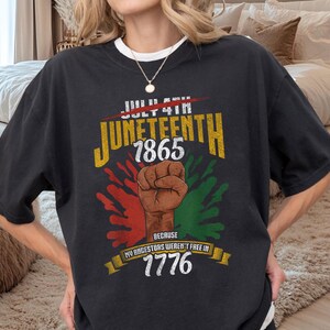 Puede incluir: Camiseta negra con cuello blanco con un gráfico de Juneteenth. El diseño incluye el texto "Juneteenth 1865" en amarillo y verde, un puño en alto y el texto "Because my ancestors weren't free in 1776."