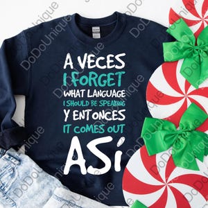 May include: Navy blue sweatshirt with the text "A Veces I Forget What Language I Should Be Speaking Y Entonces It Comes Out Asi" in white and teal. The sweatshirt is next to denim jeans and holiday decorations.