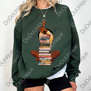 May include: Dark green sweatshirt featuring an illustration of a person sitting cross-legged, supporting a tall stack of books. The person is reaching up with one arm. The sweatshirt has a white undershirt peeking out from the bottom.