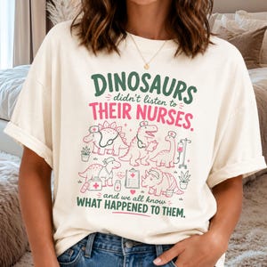 Op de afbeelding: Een off-white t-shirt met een afbeelding van verschillende dinosaurussen verkleed als medisch personeel met stethoscopen en grafieken. De tekst luidt: DINOSAURS didn't listen to THEIR NURSES. and we all know WHAT HAPPENED TO THEM. in groen en roze.