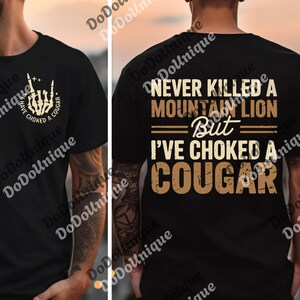 a black t-shirt with a graphic design on the back. The design includes text and a graphic of a cougar, which is a type of wild cat. The text on the t-shirt reads "NEVER KILLED A MOUNTAIN LION BUT I'VE CHOKED A COUGAR".