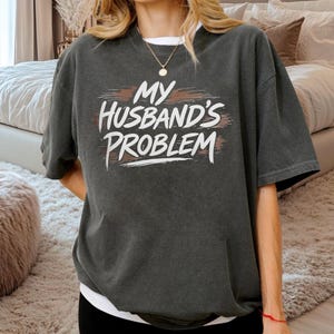 May include: Dark gray t-shirt with the text "MY HUSBAND'S PROBLEM" in white, with a brown accent. The shirt has short sleeves and a relaxed fit. The model is wearing a white undershirt.