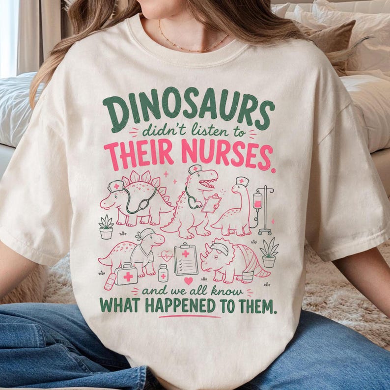 Op de afbeelding: Een cr&egrave;mekleurig t-shirt met een grafische print en groene en roze tekst die luidt: DINOSAURS didn't listen to THEIR NURSES. and we all know WHAT HAPPENED TO THEM. Het ontwerp bevat illustraties van dinosaurussen met verpleegsterskapjes.