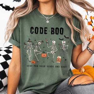 Może przedstawiać: Ciemnooliwkowy t-shirt z grafiką przedstawiającą cztery szkielety przebrane za personel medyczny w czepkach pielęgniarskich i z apteczką. Napis głosi CODE BOO oraz HERE FOR YOUR SCARE AND CARE, w towarzystwie pajęczyn, nietoperzy i dyni na Halloween.