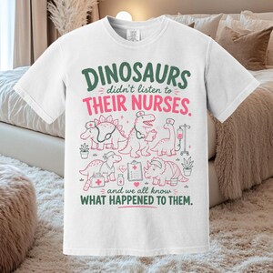 Op de afbeelding: Een wit T-shirt met korte mouwen en een afbeelding van vijf dinosaurussen verkleed als verpleegkundigen met medische hulpmiddelen. De tekst luidt: DINOSAURS didn't listen to THEIR NURSES. and we all know WHAT HAPPENED TO THEM. Groene en roze opdruk.