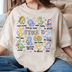 以下が含まれることがあります： クリーム色のTシャツで、様々な漫画の恐竜のカラフルなグラフィックと「IT'S OKAY TO」というテキストが描かれています。また、「need routines」、「avoid eye contact」、「make mistake」、「be yourself」、「need space」、「take breaks」、「feel deeply」、「unmask」、「communicate differently」、「be different」、「stim」というフレーズも含まれています。