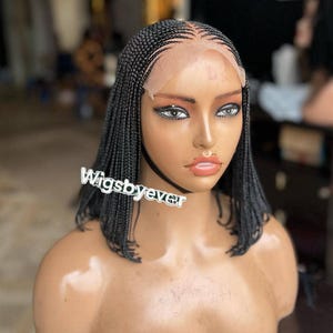 Pode incluir: Uma peruca preta trançada em uma cabeça de manequim. A peruca apresenta pequenas tranças e uma frente de renda. O texto "Wigsbyever" é visível na imagem. A peruca é estilizada em um corte bob.