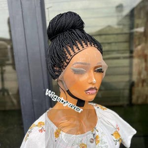 May include: A black braided wig styled in a high bun. The wig is on a mannequin head with a transparent lace front. The mannequin is wearing a white top with a floral pattern. The text "Wigsbyever" is visible.