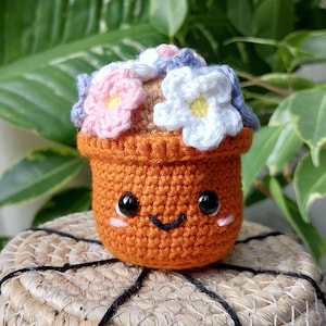Op de afbeelding: Een gehaakte bloempot met een glimlachend gezicht en roze, witte en paarse bloemen. De pot is oranje en staat op een geweven mand.