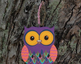 May include: A purple owl ornament with yellow eyes, orange and pink accents, and a colorful feathered pattern on its chest. The owl is hanging from a string.
