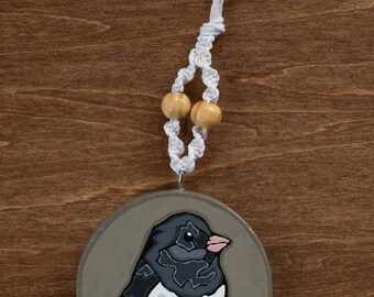 May include: A round, wooden ornament featuring a painted bird with black, white, and gray feathers, a pink beak, and brown feet. The ornament is suspended by a white macrame cord with two wooden beads. The background is a brown wooden surface.