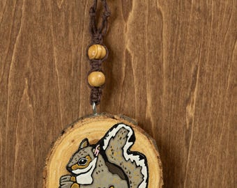 May include: A hand-painted wooden ornament featuring a gray squirrel holding a nut. The squirrel is painted on a round wood slice with a natural bark edge. The ornament hangs from a brown macrame cord with two wooden beads.
