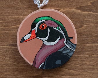 May include: A round, wooden pendant featuring a hand-painted wood duck. The duck has a green head, black and white markings, and a red eye and beak. The pendant is attached to a white, braided cord.