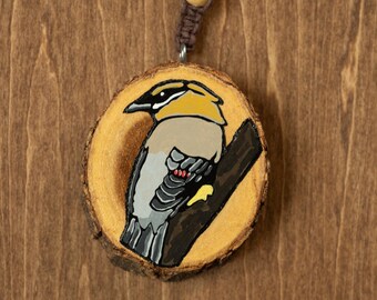 May include: A round, wooden ornament featuring a hand-painted bird perched on a branch. The bird has yellow, black, gray, and white feathers. The ornament is attached to a brown cord with a wooden bead.