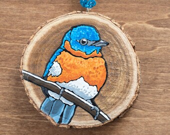 May include: Hand-painted wooden ornament featuring a bluebird perched on a branch. The bird has a blue head and wings, an orange breast, and white accents. The ornament is round, made of a wood slice, and has a blue string for hanging.