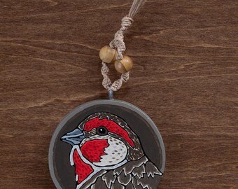 May include: A round, hand-painted wooden ornament featuring a detailed bird portrait. The bird has a red head and chest, with brown and white accents. It hangs from a natural twine cord with two wooden beads. The background is a dark brown wood grain.