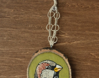 May include: A hand-painted wooden ornament featuring a sparrow with a detailed design. The ornament is painted in shades of brown, gray, orange, and yellow. The sparrow is perched on a green background. The ornament is attached to a macrame cord.