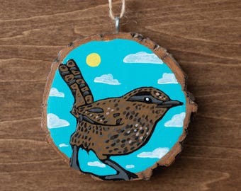 May include: Hand-painted wooden ornament featuring a brown bird with a speckled pattern, set against a blue sky with white clouds and a yellow sun. The bird is perched on a wooden slice, with a natural wood grain edge and a twine loop for hanging.
