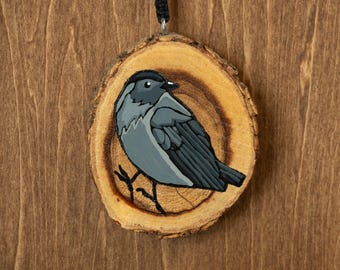 May include: A circular wood slice ornament featuring a hand-painted illustration of a small bird with grey and black plumage. The bird is depicted in profile, facing right, with a black cap and dark wings. A black cord is attached to the top.
