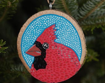 May include: A hand-painted wooden ornament featuring a red cardinal with a black head and beak against a blue background with white dots.