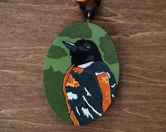 May include: Oval-shaped wooden pendant featuring a painted bird. The bird has black feathers, orange accents, and a white chest, set against a green background with camouflage-like patterns. The pendant hangs from a black cord with orange beads.