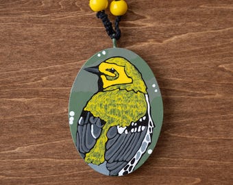 May include: Oval-shaped pendant featuring a hand-painted bird design. The bird has yellow and green plumage, a black beak, and gray wings. The pendant is suspended from a black cord with yellow beads.