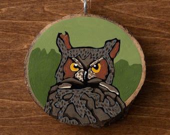 May include: Hand-painted wooden owl ornament. The owl has yellow eyes, brown and gray feathers, and is set against a green background. The ornament is round and has a natural wood grain edge, with a twine loop for hanging.