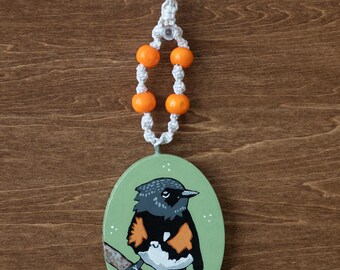 May include: An oval-shaped, hand-painted wooden ornament featuring a bird perched on a branch. The bird has black, gray, orange, and white plumage against a light green background. The ornament is suspended by a white cord with orange beads.