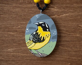 May include: Oval pendant necklace featuring a hand-painted bird design. The bird has yellow, black, and white markings, set against a gray and green background. The necklace has a black cord and yellow beads.