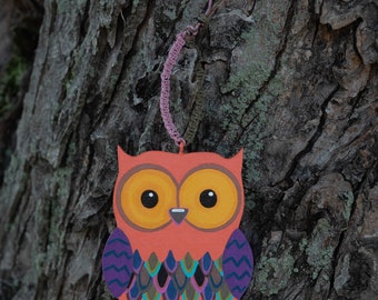 May include: A colorful wooden owl ornament with orange, purple, and green paint. The owl has large yellow eyes and a detailed pattern on its body. The ornament is hanging from a pink string.