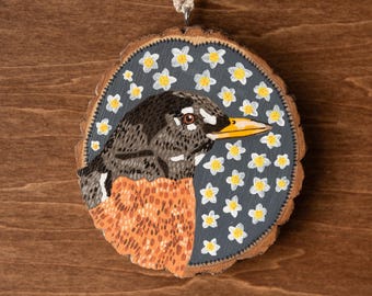 May include: A hand-painted wooden ornament featuring a robin with an orange breast and a gray head, surrounded by white flowers on a dark blue background.