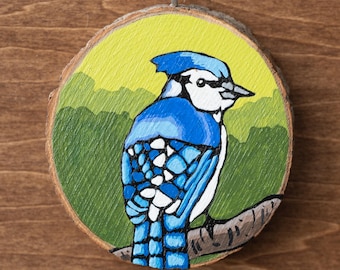 May include: Hand-painted wooden ornament featuring a blue jay perched on a branch. The bird is depicted with shades of blue, white, and black against a yellow and green background. The ornament is round, with a natural wood edge and a string for hanging.