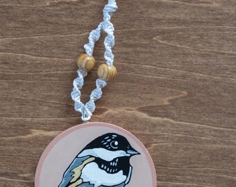 May include: Round wooden ornament featuring a painted bird perched on a branch. The bird has black, white, and gray feathers. The ornament is attached to a white cord with two wooden beads. The background is a wood grain.