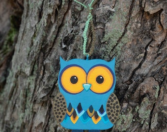 May include: A blue and yellow owl ornament with black and teal accents hanging from a tree branch. The owl has large, round eyes and a small beak.