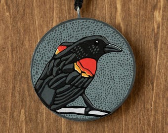 May include: A round, painted wooden pendant featuring a black bird with red and yellow markings on a gray background. The bird is perched on a white line, and the pendant hangs from a black cord.