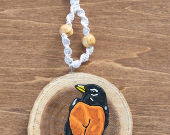 May include: A round wooden ornament featuring a hand-painted robin. The bird has a black head and wings, an orange breast, and a yellow beak. It hangs from a white macrame cord with two wooden beads. The ornament is approximately 3 inches in diameter.