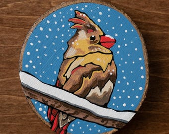May include: Hand-painted wooden ornament featuring a cardinal bird perched on a snow-covered branch. The bird has red, yellow, and brown feathers against a blue background with white snow dots. A metal hook and twine are attached for hanging.