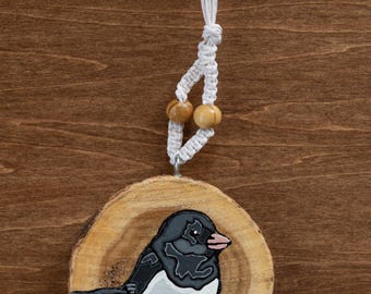 May include: A hand-painted wooden ornament featuring a black and white bird with a pink beak. The bird is perched on a brown surface, set against a natural wood slice. The ornament hangs from a white macrame cord with two wooden beads.