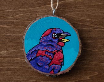 May include: A round wooden ornament featuring a painted bird. The bird has purple and red feathers, a yellow beak, and a black eye, set against a turquoise background. A natural twine loop is attached for hanging.