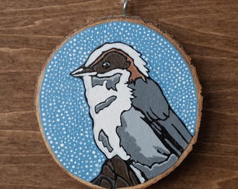 May include: A hand-painted wooden ornament featuring a bird illustration. The bird has gray, white, and brown feathers, set against a light blue background with white dots. The ornament is round, with a natural wood edge and a twine loop for hanging.