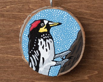 May include: Hand-painted wooden ornament featuring a woodpecker. The bird has a red head, black and white body, and yellow beak, set against a blue background with white dots. The ornament is round, made from a wood slice, and has a natural wood grain edge.