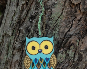 May include: A blue and gold owl ornament with a green string hanging from the top. The owl has large yellow eyes and a detailed design on its feathers.