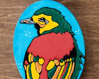 May include: Hand-painted wooden pendant featuring a colorful bird design. The oval pendant has a blue background with white dots. The bird has green, yellow, and red details. The pendant is attached to a red woven cord necklace.