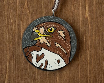 May include: A circular pendant featuring a hand-painted illustration of a hawk with brown and white feathers, a yellow eye, and a dark beak. The hawk is set against a dark gray background with small white dots. A braided cord is attached to the top.