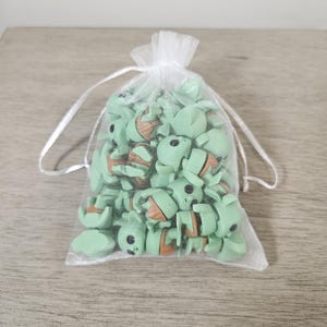 Puede incluir: Una bolsa transparente blanca con cord&oacute;n llena de peque&ntilde;as figuras verdes y marrones. Las figuras se asemejan a un personaje de una popular serie de ciencia ficci&oacute;n. Tienen ojos negros y detalles en naranja. La bolsa est&aacute; sobre una superficie de madera clara.