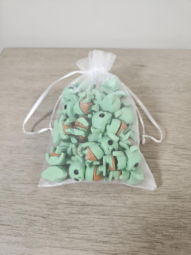 Puede incluir: Una bolsa transparente blanca con cord&oacute;n llena de peque&ntilde;as figuras verdes y marrones. Las figuras se asemejan a un personaje de una popular serie de ciencia ficci&oacute;n. Tienen ojos negros y detalles en naranja. La bolsa est&aacute; sobre una superficie de madera clara.