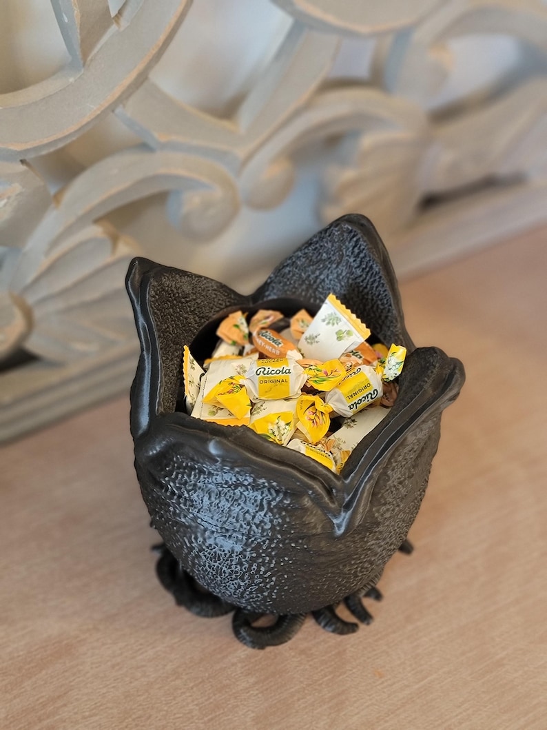 May include: A black, textured, decorative bowl filled with individually wrapped Ricola candies. The bowl has a unique, organic shape with a wide opening and a textured exterior. The candies are yellow and white.
