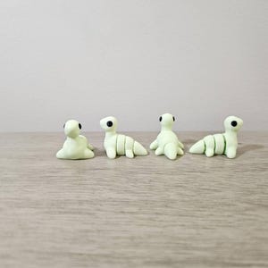 May include: Four small, light green clay dinosaur figurines with black eyes are arranged on a wooden surface. The dinosaurs are in various poses, some sitting and others standing. The figurines are handmade.
